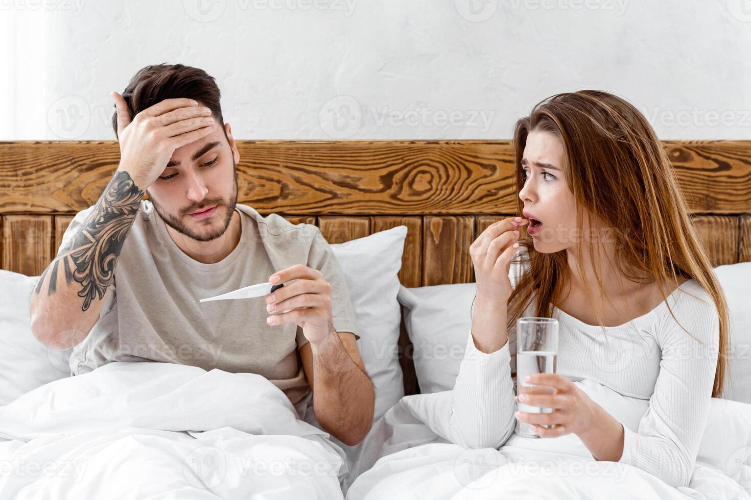 Treated together in bed. Sad woman drinking pill, holding glass of water and looking at husband, man touches forehead and looks at thermometer, panorama photo