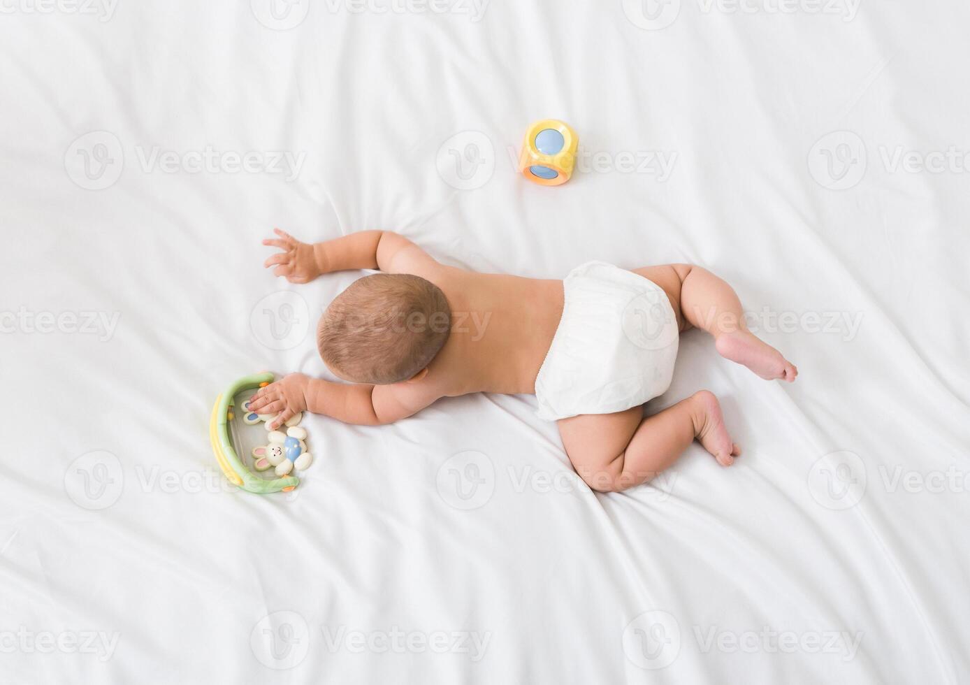 Small baby in diaper lying on his belly on white bed with toys, top view, copy space photo