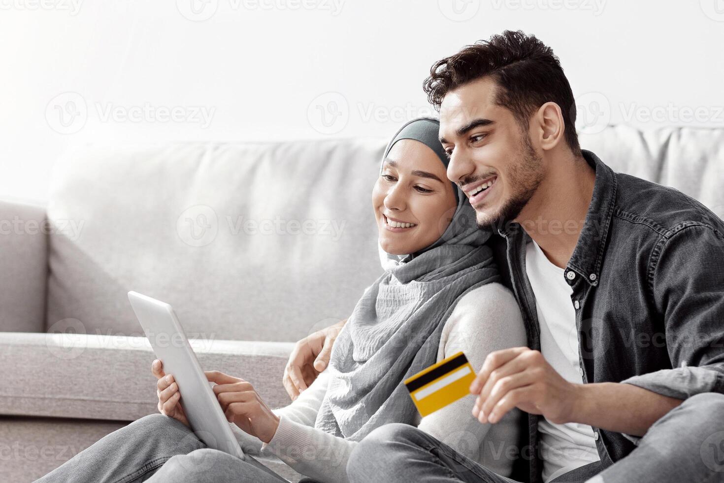 Happy young arabic couple husband and wife in hijab with digital tablet and credit card sitting on floor among paper boxes in new apartment, buying furniture for their house online, copy space photo