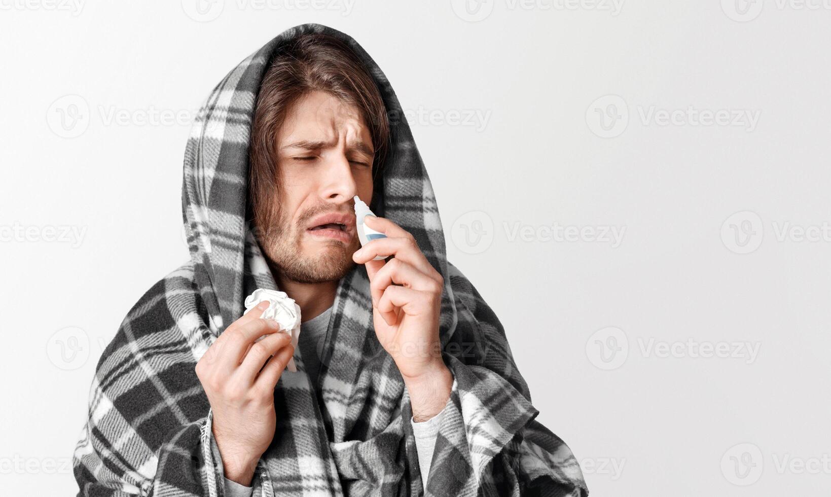 Virus and cold. Man in blanket drips into his nose and holds napkin in his hand, face expresses suffering, isolated on gray background photo