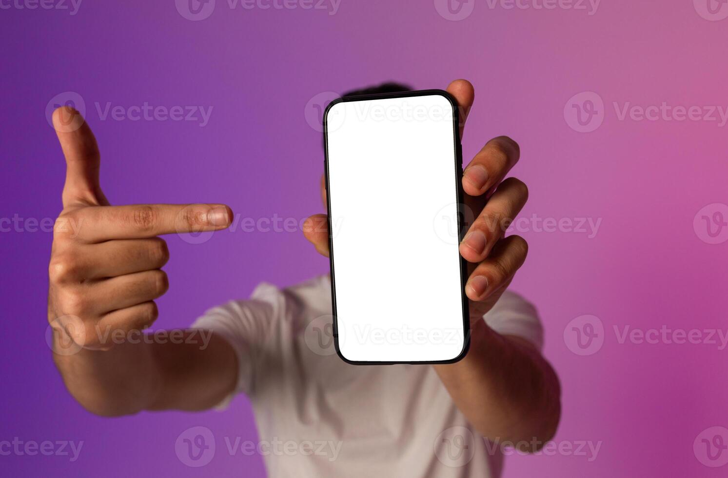 Unrecognizable young man holding smartphone in front of his face, pointing at empty screen, offering space for mobile app or website, demonstrating mockup for online ad in neon light, selective focus photo