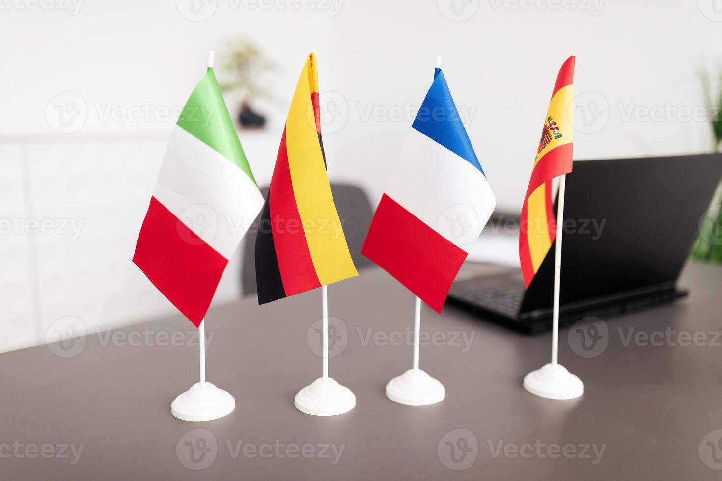 Four small national flags, including Italy, Germany, France, and Spain, are arranged on a desk in a contemporary office. The workspace features a laptop and a clean design, reflecting professionalism. photo