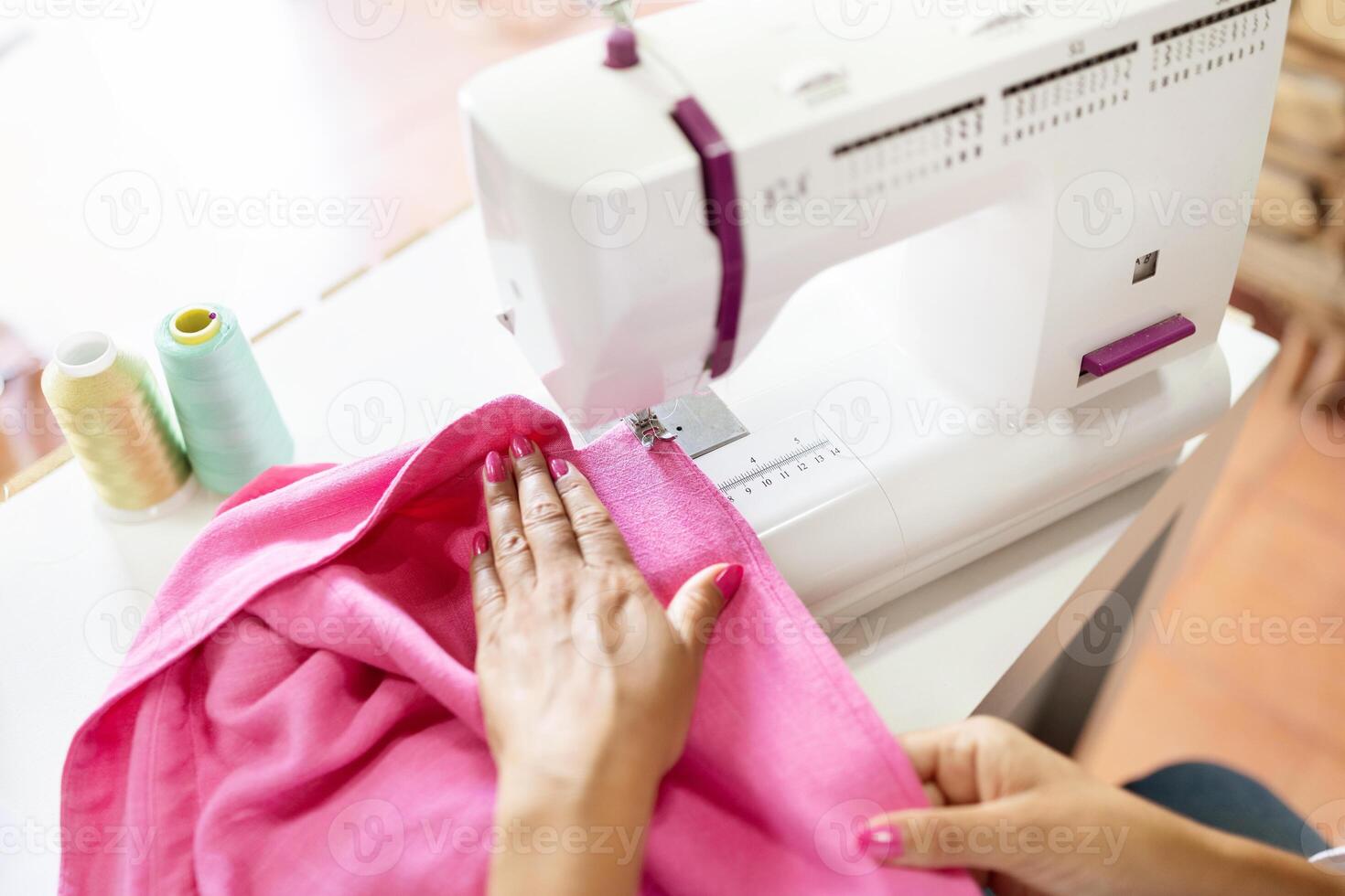 A mature Hispanic woman skillfully works on a sewing machine, focusing on vibrant pink fabric. Colorful spools of thread are visible nearby, showcasing her creativity and dedication as a designer. photo
