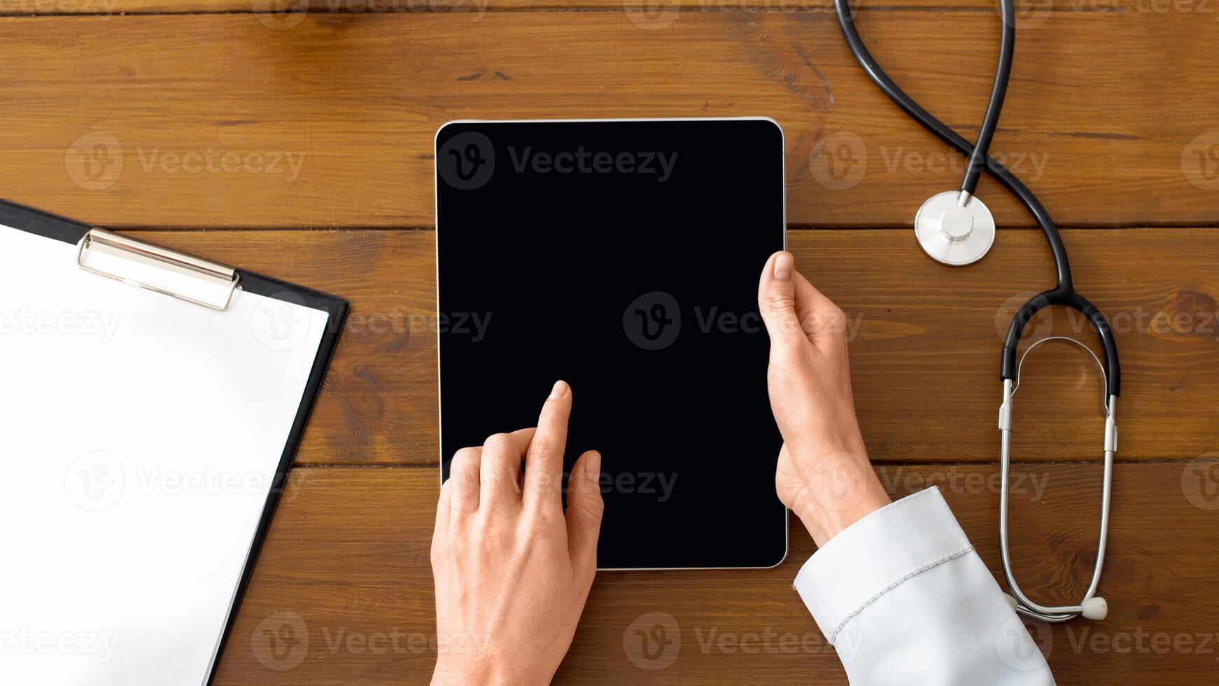 Modern private practice of doctor and patient visit. Hands of female therapist in white coat works on tablet with blank screen on wooden table with stethoscope, apple, plant and glasses, top view, pov photo