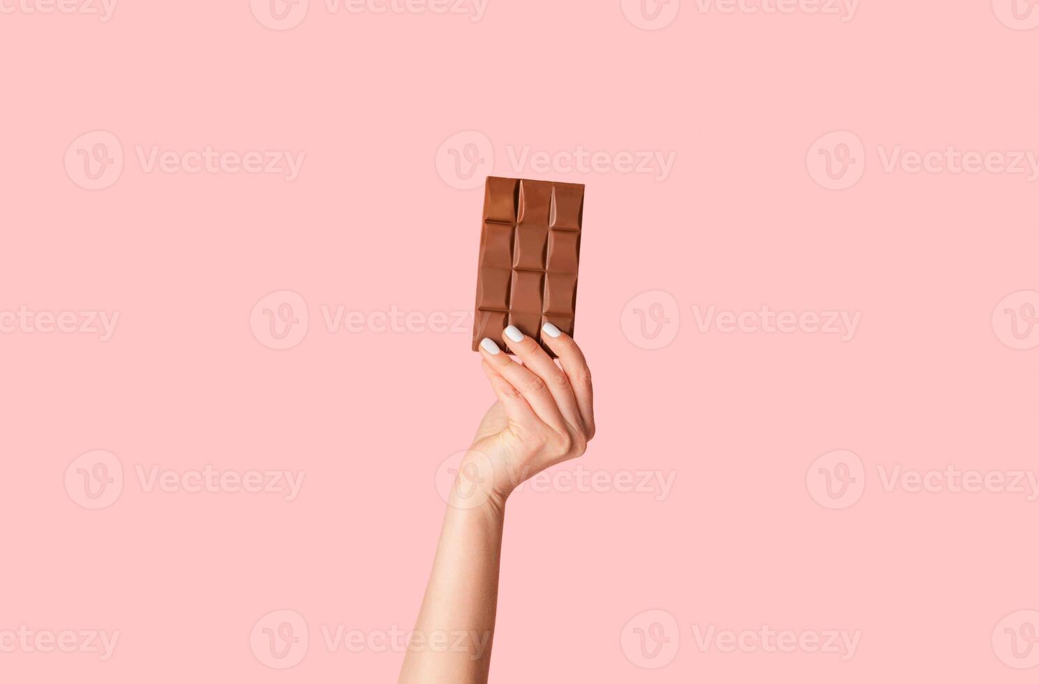 Millennial woman holding bar of organic chocolate over pink background, close up of hand. Panorama photo