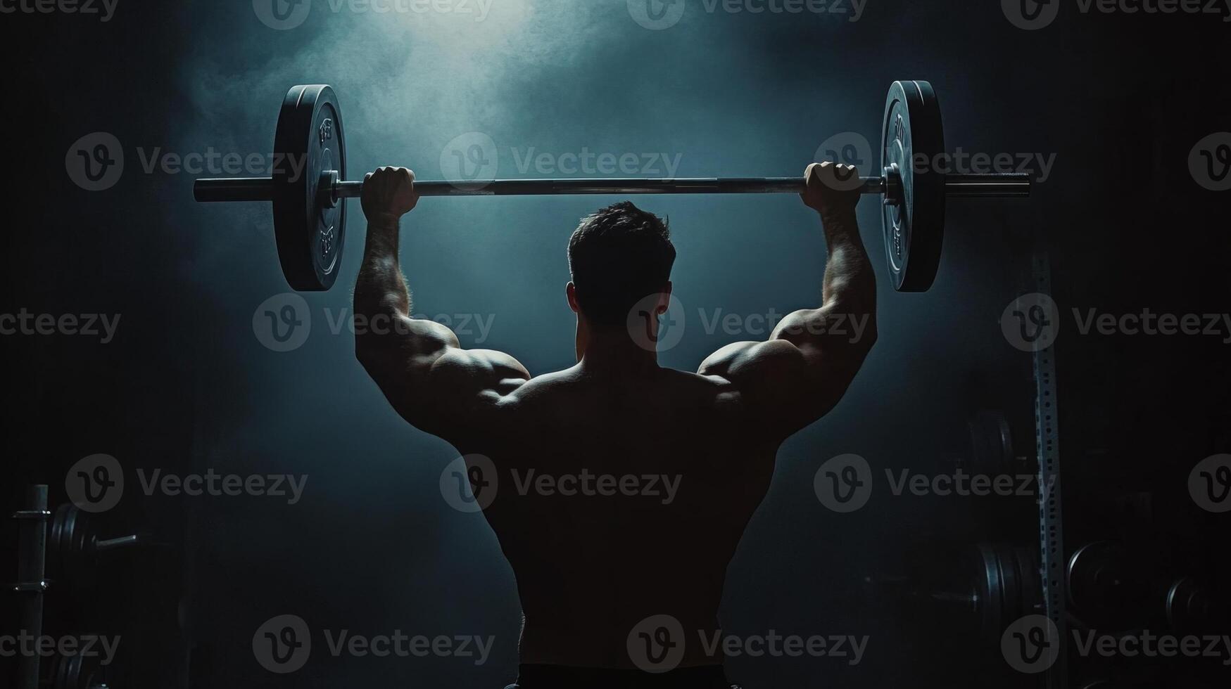 A man with a muscular back lifts a heavy barbell in a dimly lit gym. He is facing away from the camera and his arms are extended above his head as he lifts the weight. photo