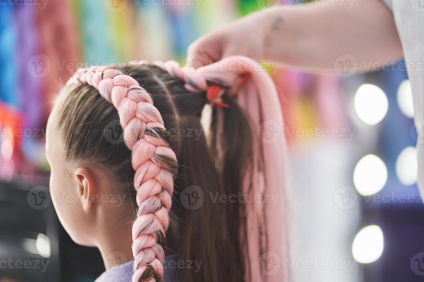 The master braids the child's pigtails from a pink kanekalon. Close-up, side view. The hands of the master weave the kanekalon material into the hair photo