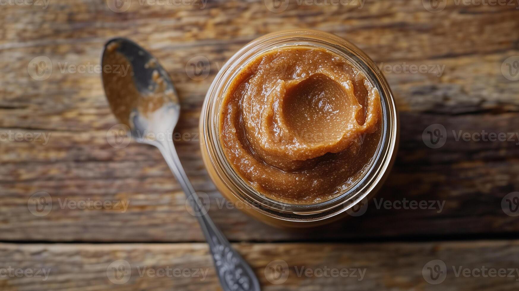 Glass jar with thick caramel-colored spread on a wooden surface with a spoon, food photography photo