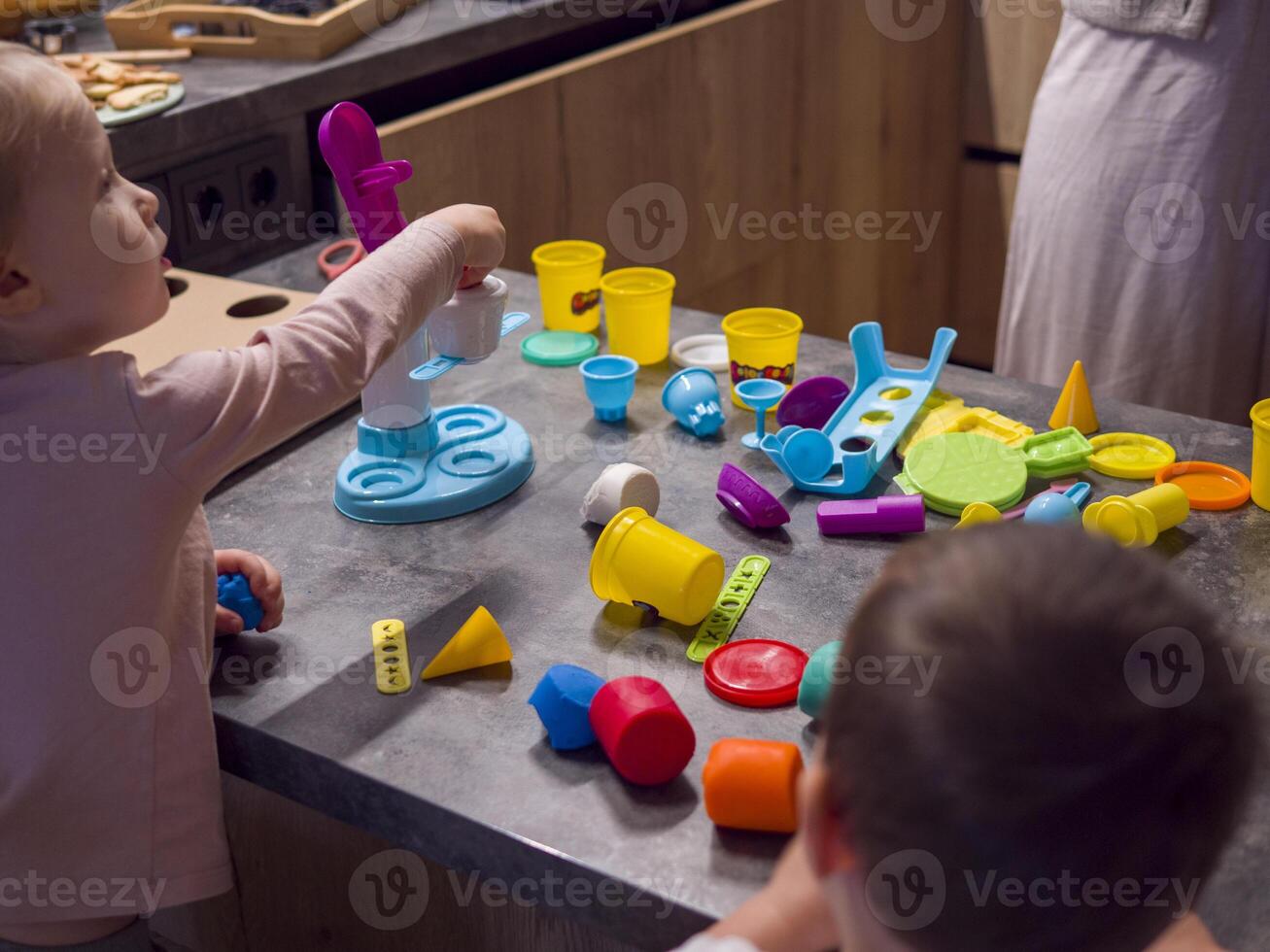 Two children enjoy creative playtime in kitchen using vibrant modeling clay and toys, while an adult observes. toys add fun making this educational activity. Concept of teaching children photo