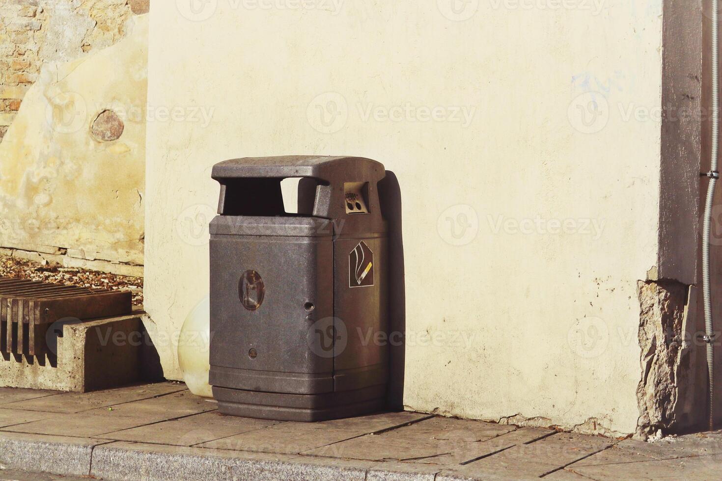 Black plastic garbage can with lid standing near the wall of a light yellow building in evening sunlight photo