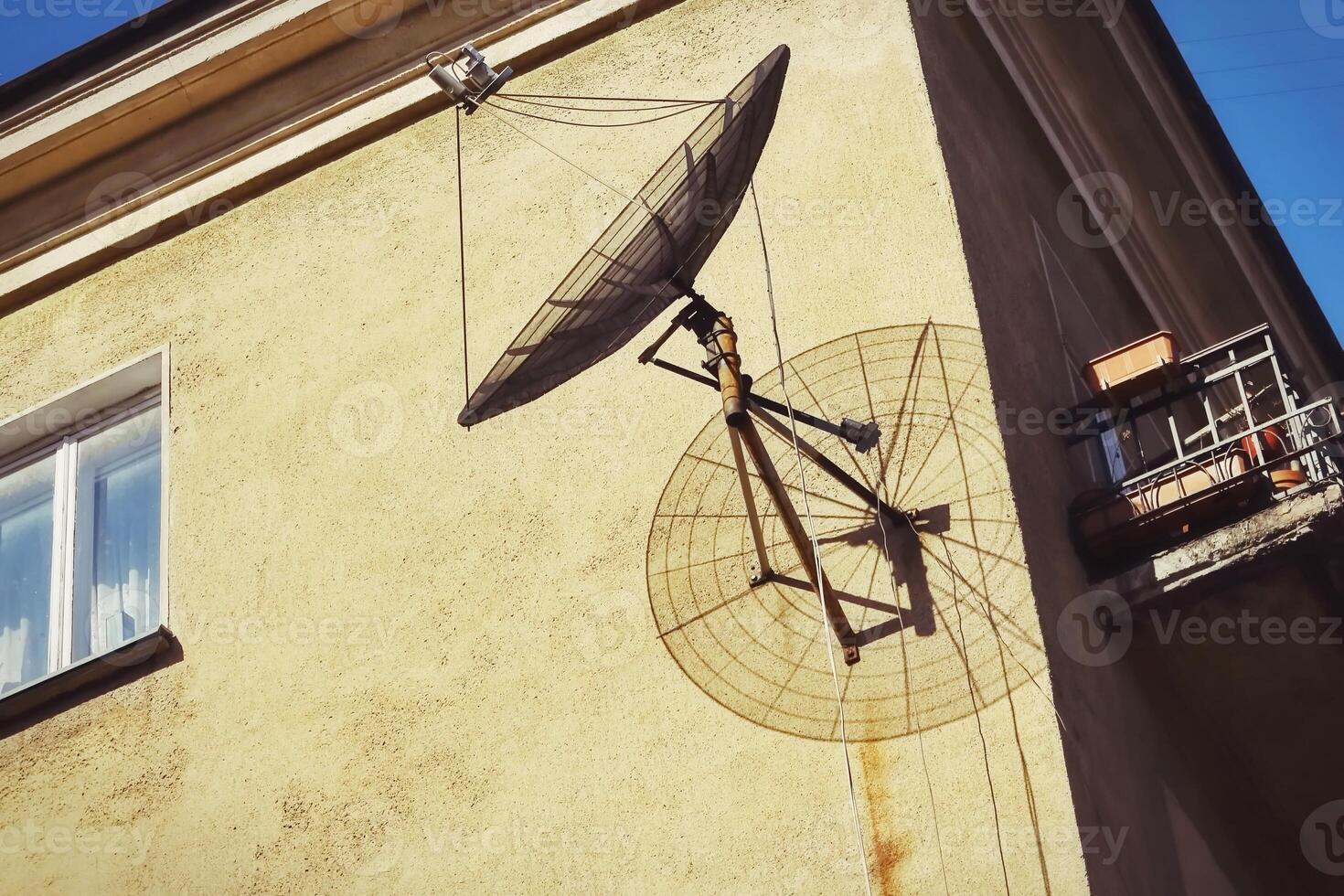A round satellite dish casting a nice shadow on the corner of a building against a yellow building wall with a blue sky above photo