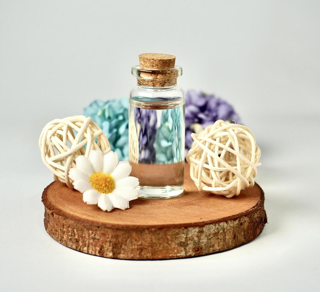 Transparent glass vial bottle container with brown cork stopper lid on wooden coaster with flowers and rattan ball decoration isolated on white background. photo