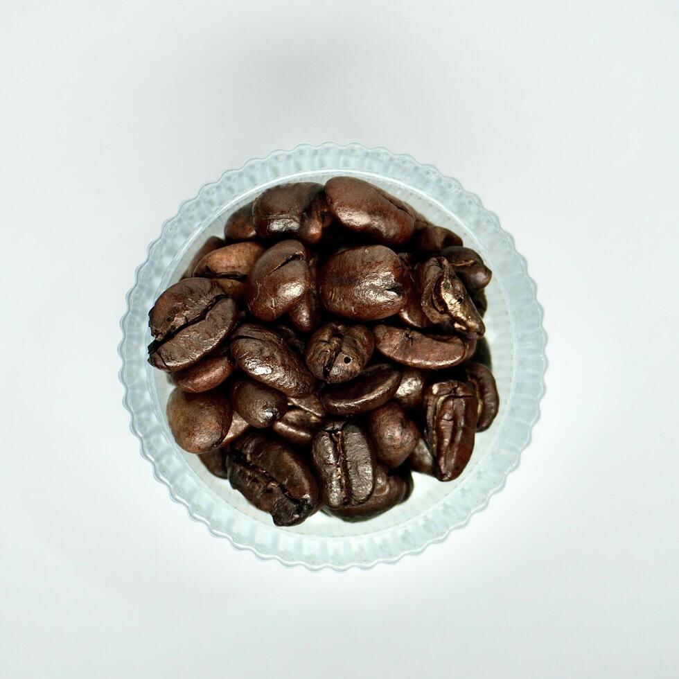 Group of brown coffee beans in round glass bowl container isolated on square white background from above angle view. photo