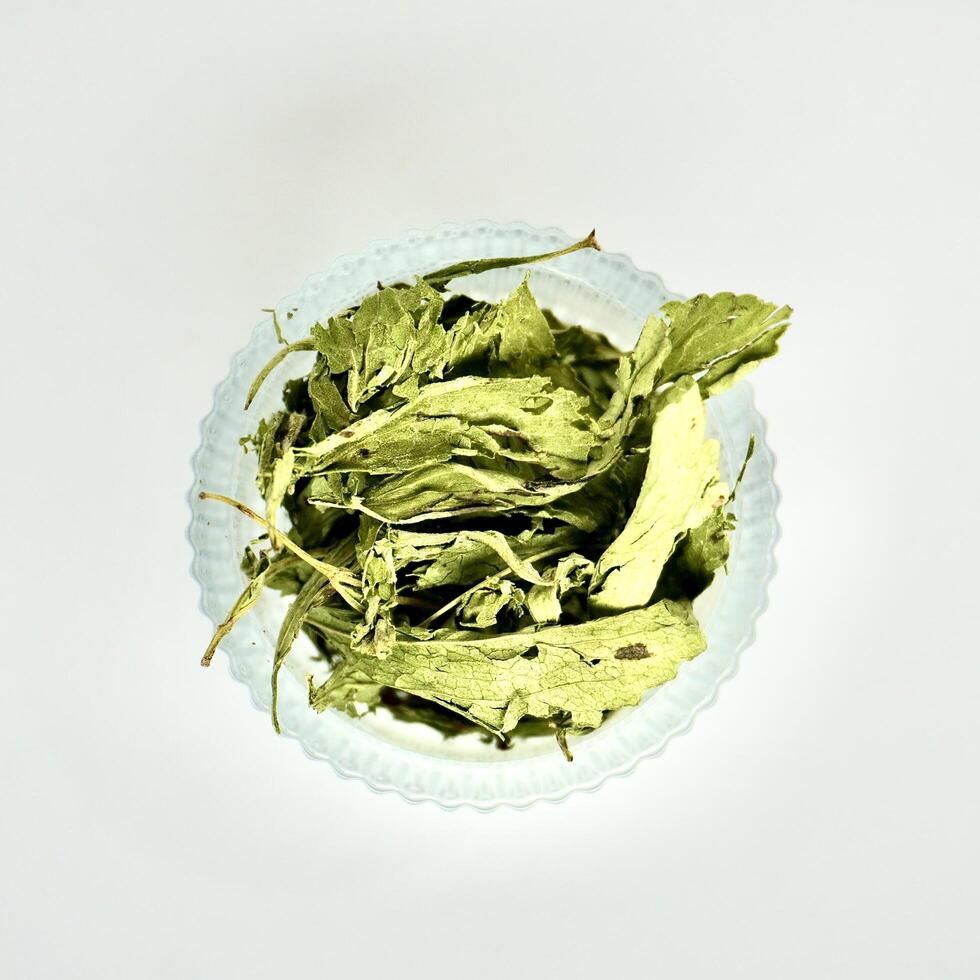 Crushed stevia plant leaves in round glass bowl isolated on square white background from above angle view. photo