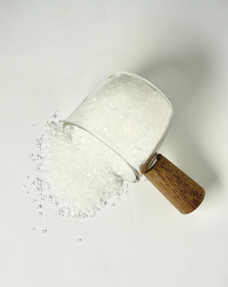 Pile of salt food flavoring ingredient spilled from glassware cup with wooden handle isolated on vertical white background from above angle view. photo