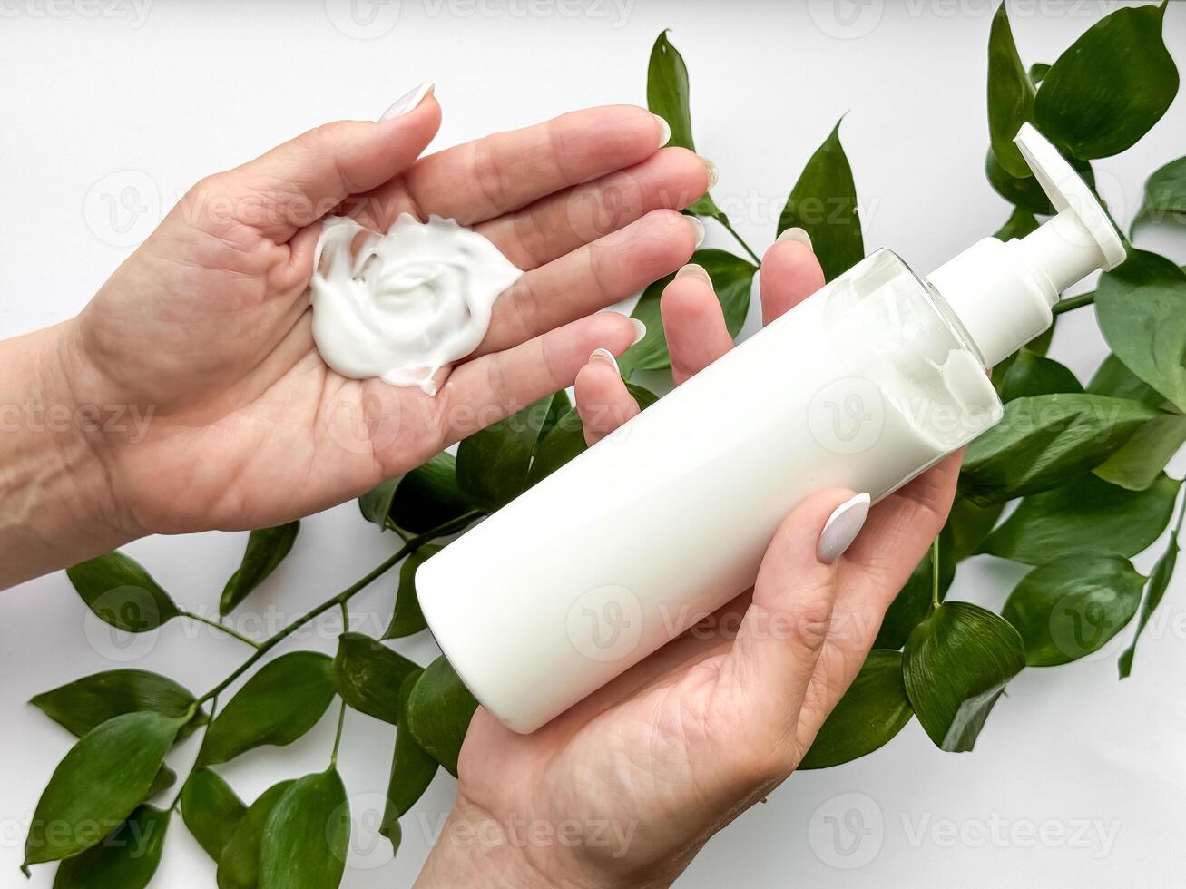 Close up of middle age woman hand holding white lotion bottle with green leaves on white background. Skincare and natural beauty concept. Healthcare, wellness, and organic product poster. photo