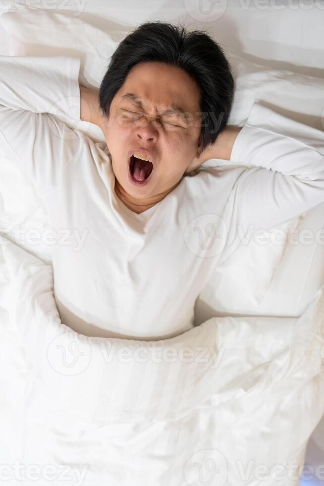 An Asian man is captured in a moment of genuine yawn as he rests on a soft white bed and pillow. The image evokes a sense of contagious yawning, inviting viewers to join in the action. photo