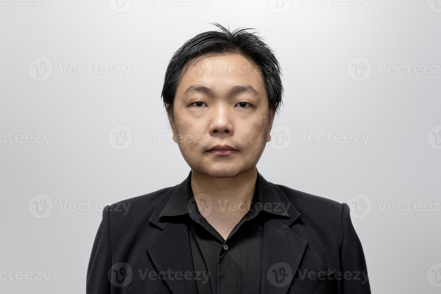 Asian man in his middle age, dressed in a stylish black suit and shirt, poses against a neutral white-gray backdrop. photo