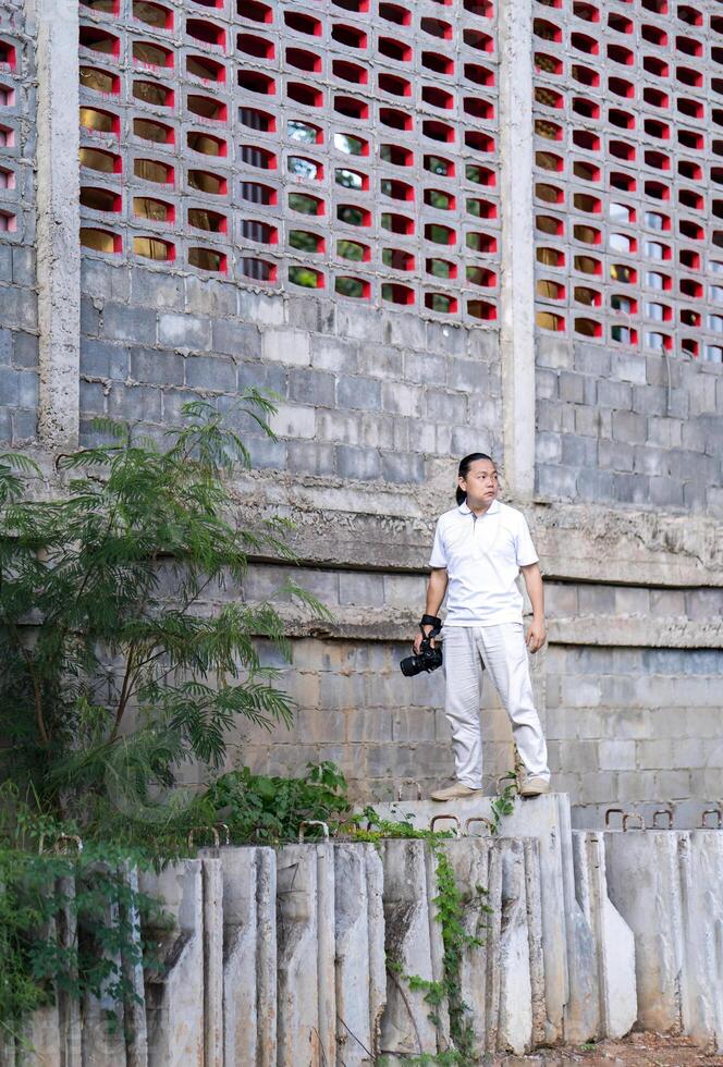 Asian Professional cameraman pretentious stands on construction, concrete barricade and looks into the sky, He thinks how to post for a photo shooting in smart image.