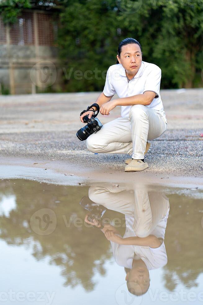 Asian man holds a big black mirrorless camera medium format type on cement asphalt ground in front of water reflex. photo
