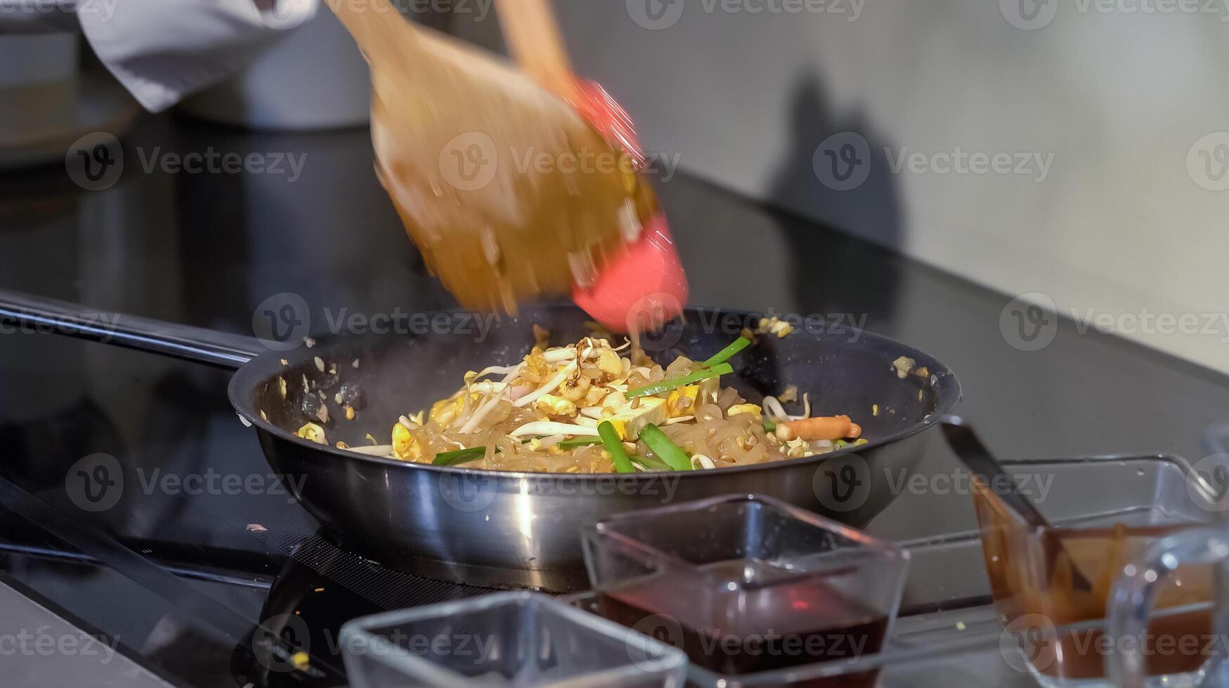 A cook stirs fry Pad Thai noodles in a pan with spatulas on electric stoves. Sauce dishes for cooking traditional Pad Thai. photo