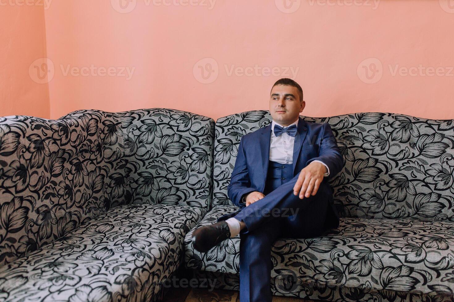 A man in a suit and bow tie sits on a couch photo