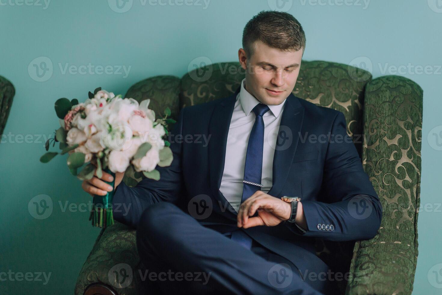 A man in a suit and tie is sitting in a green chair and looking at his watch photo