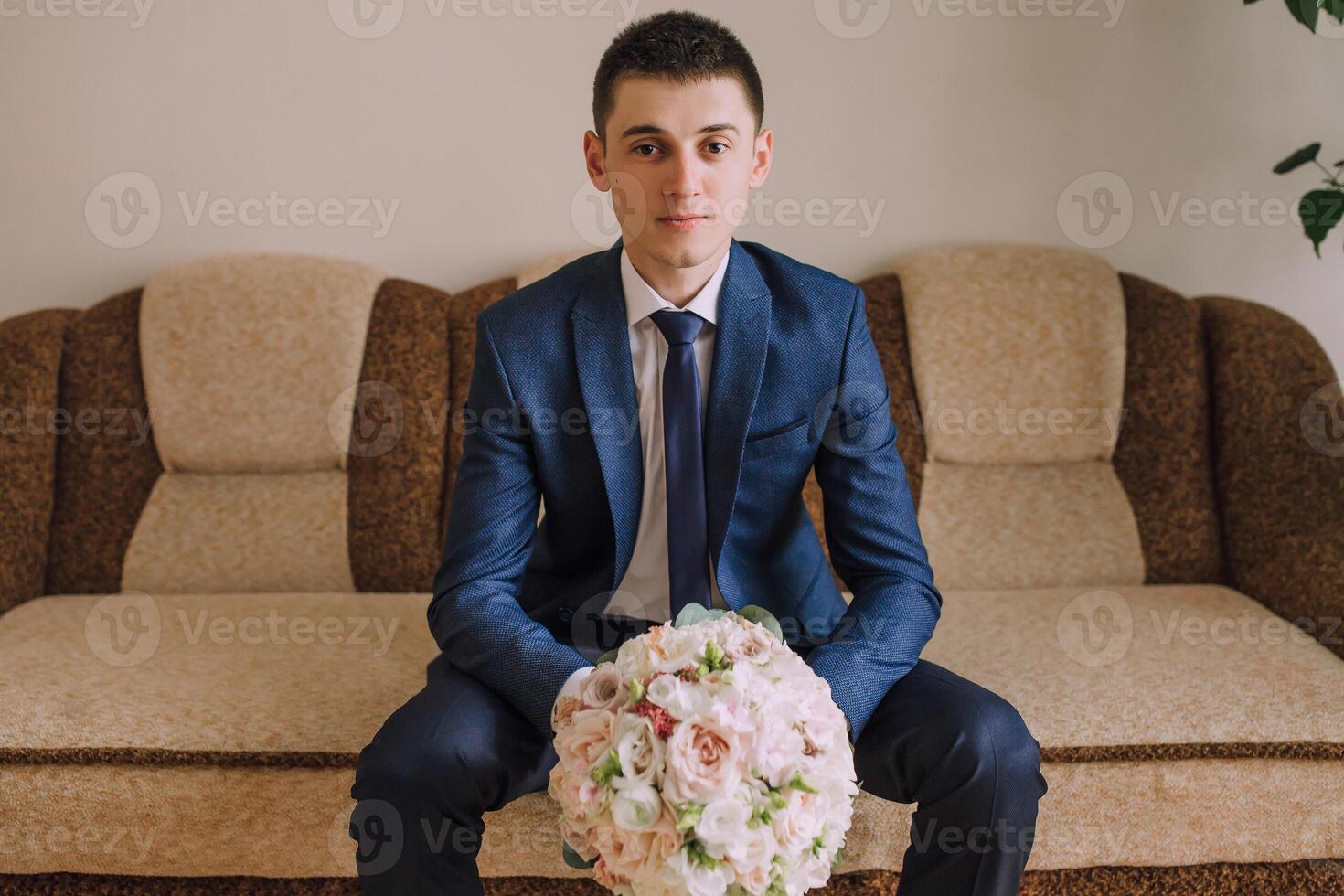A man in a suit and tie sits on a couch holding a bouquet of flowers photo