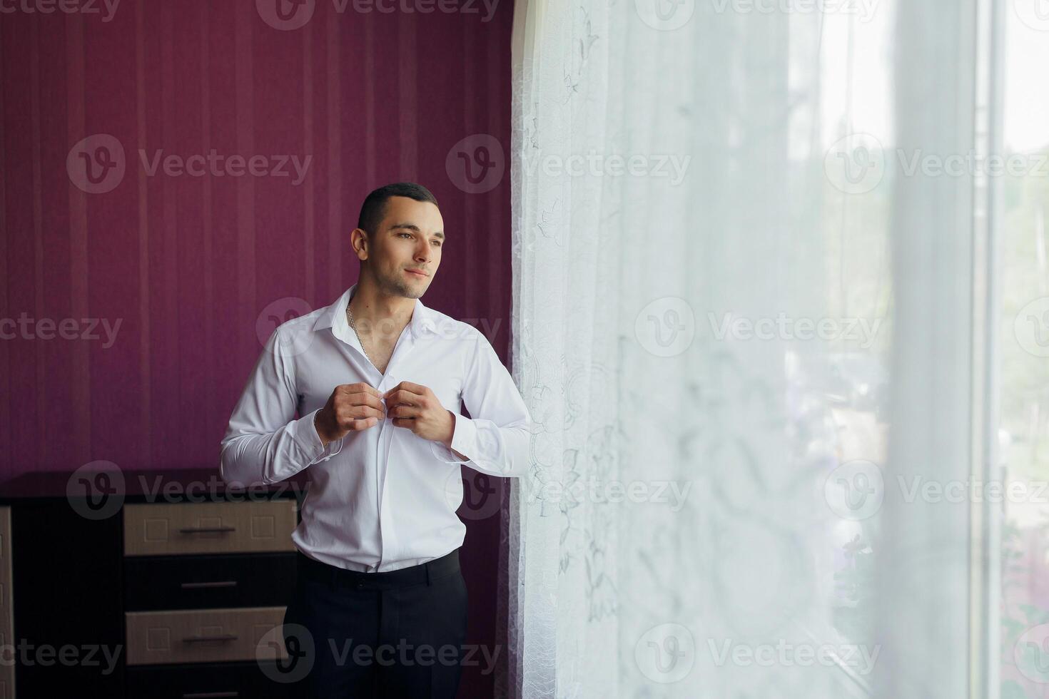 A man in a white shirt is adjusting his collar while looking out the window photo