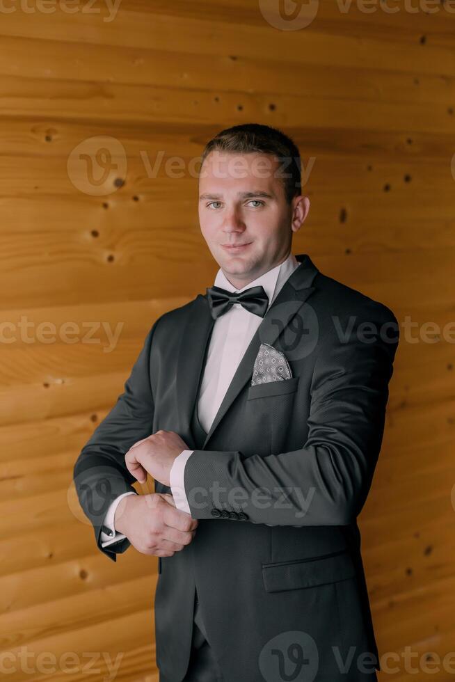 A man in a black suit and bow tie is standing in front of a wooden wall photo