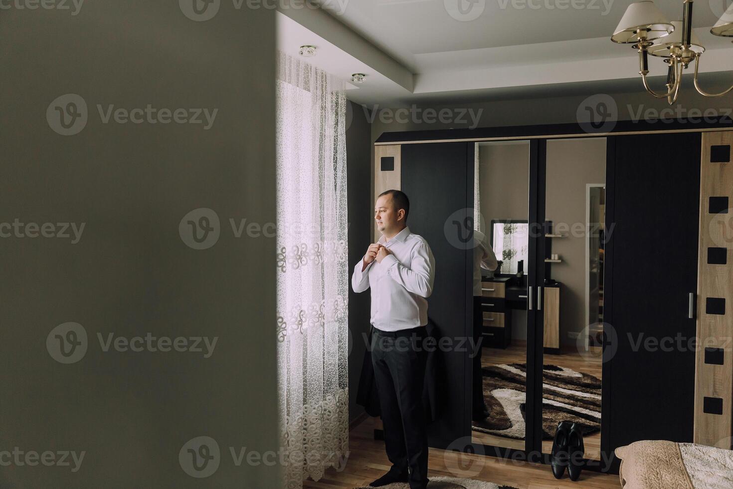 A man is getting dressed in a room with a window and a mirror photo