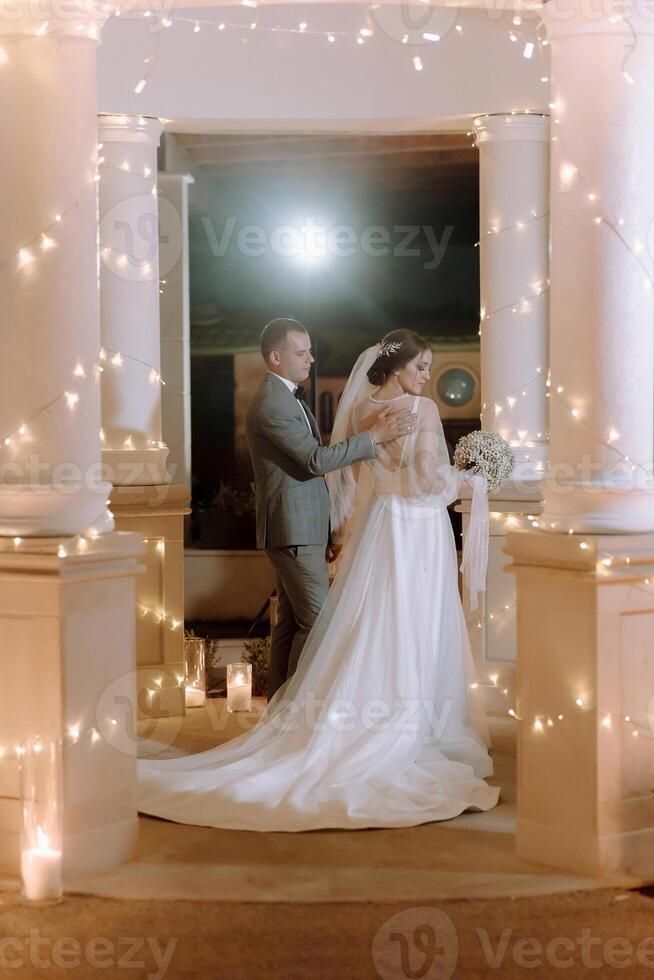 A bride and groom stand in front of a building with lights on photo