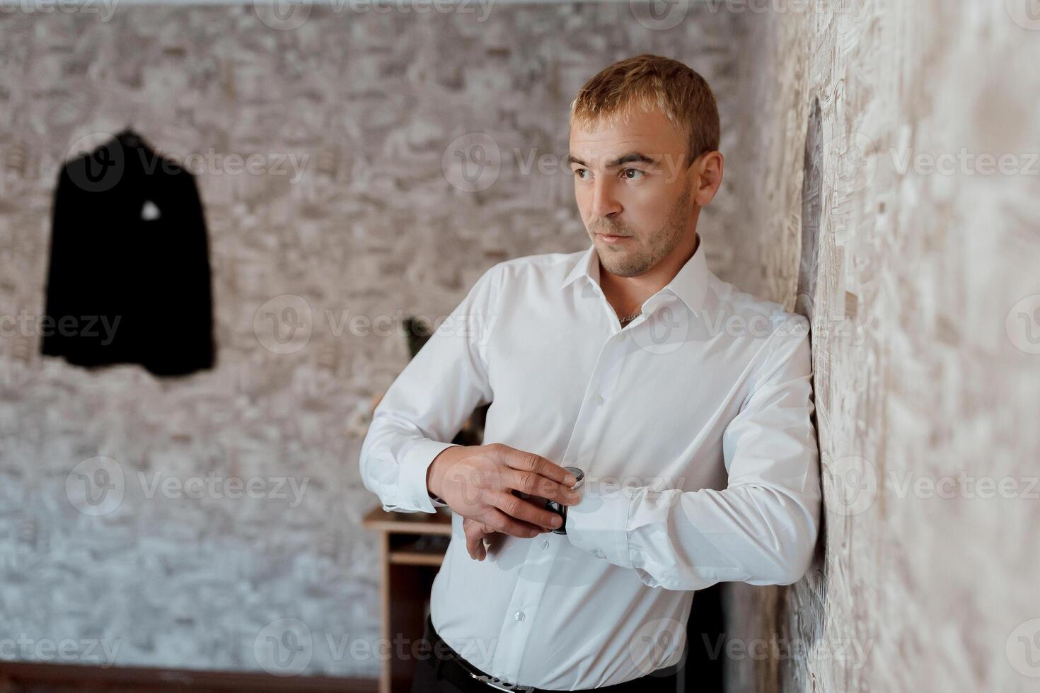 A man in a white shirt is standing in front of a wall with a black jacket hangin photo