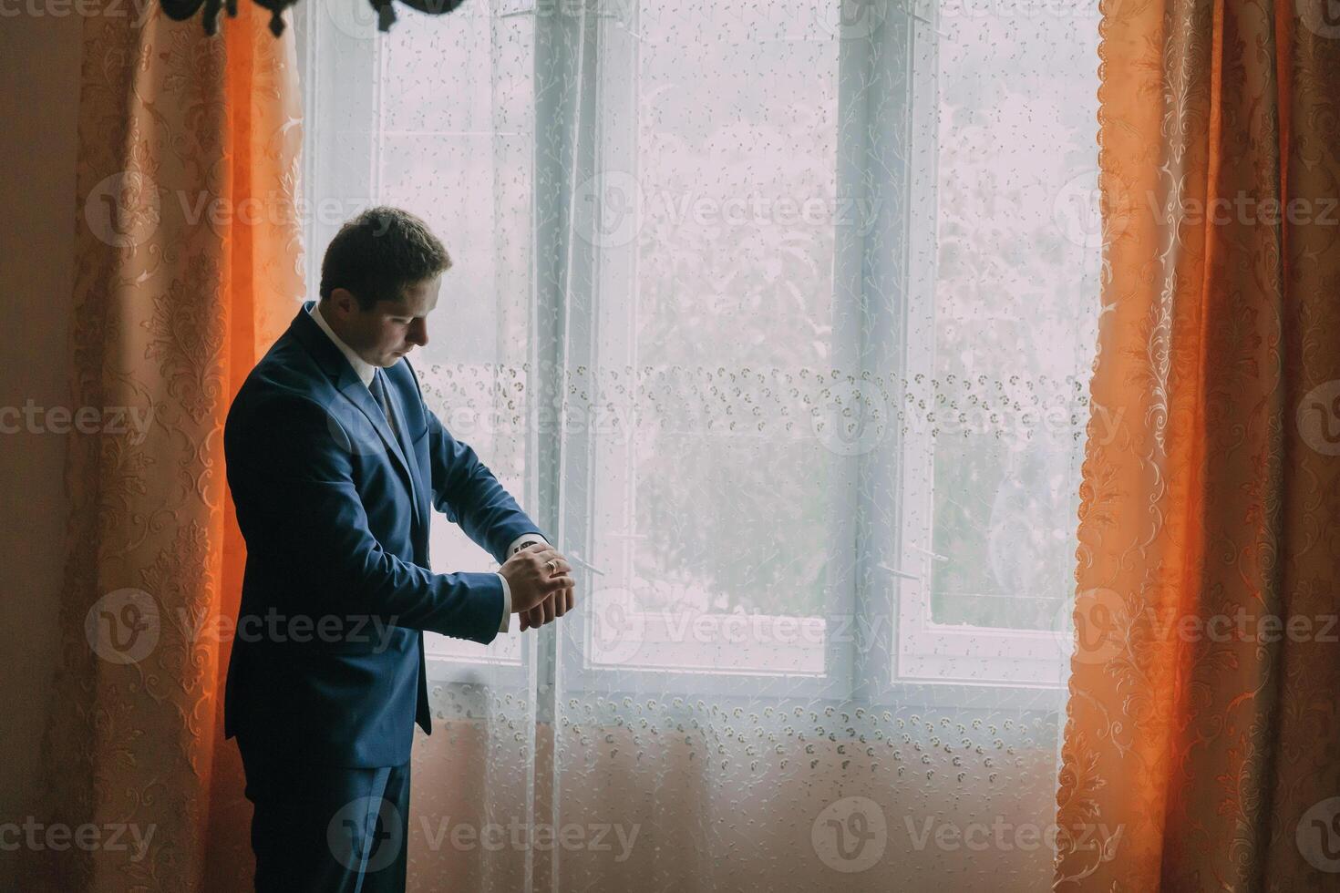 A man in a suit is standing in front of a window, looking at his watch photo
