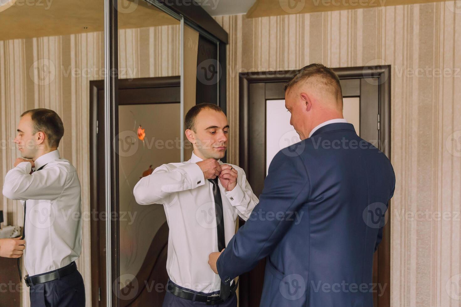 Two men are getting dressed up, one of them is wearing a blue suit photo