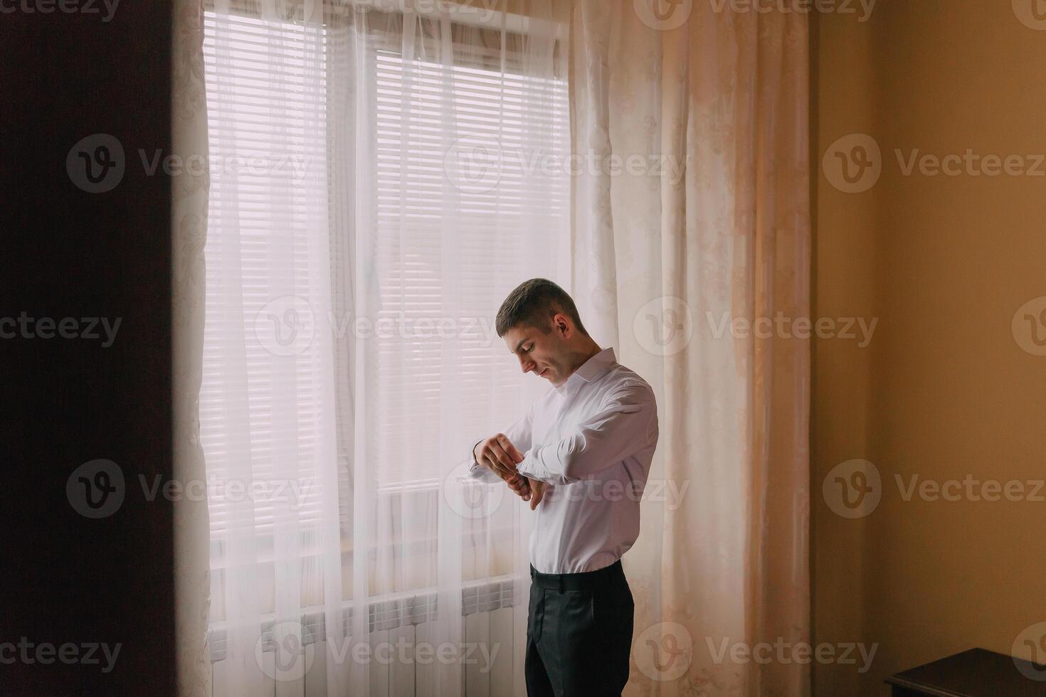 A man is standing in front of a window, wearing a white shirt and black pants photo