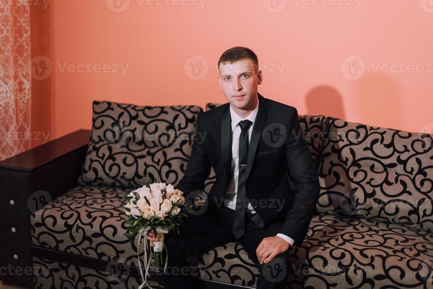 A man in a suit and tie sits on a couch holding a bouquet of flowers photo