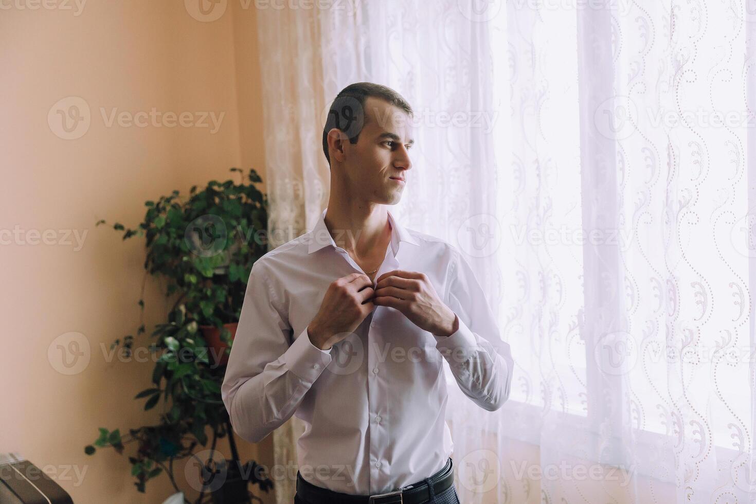 A man in a white shirt is standing in front of a window, adjusting his collar photo