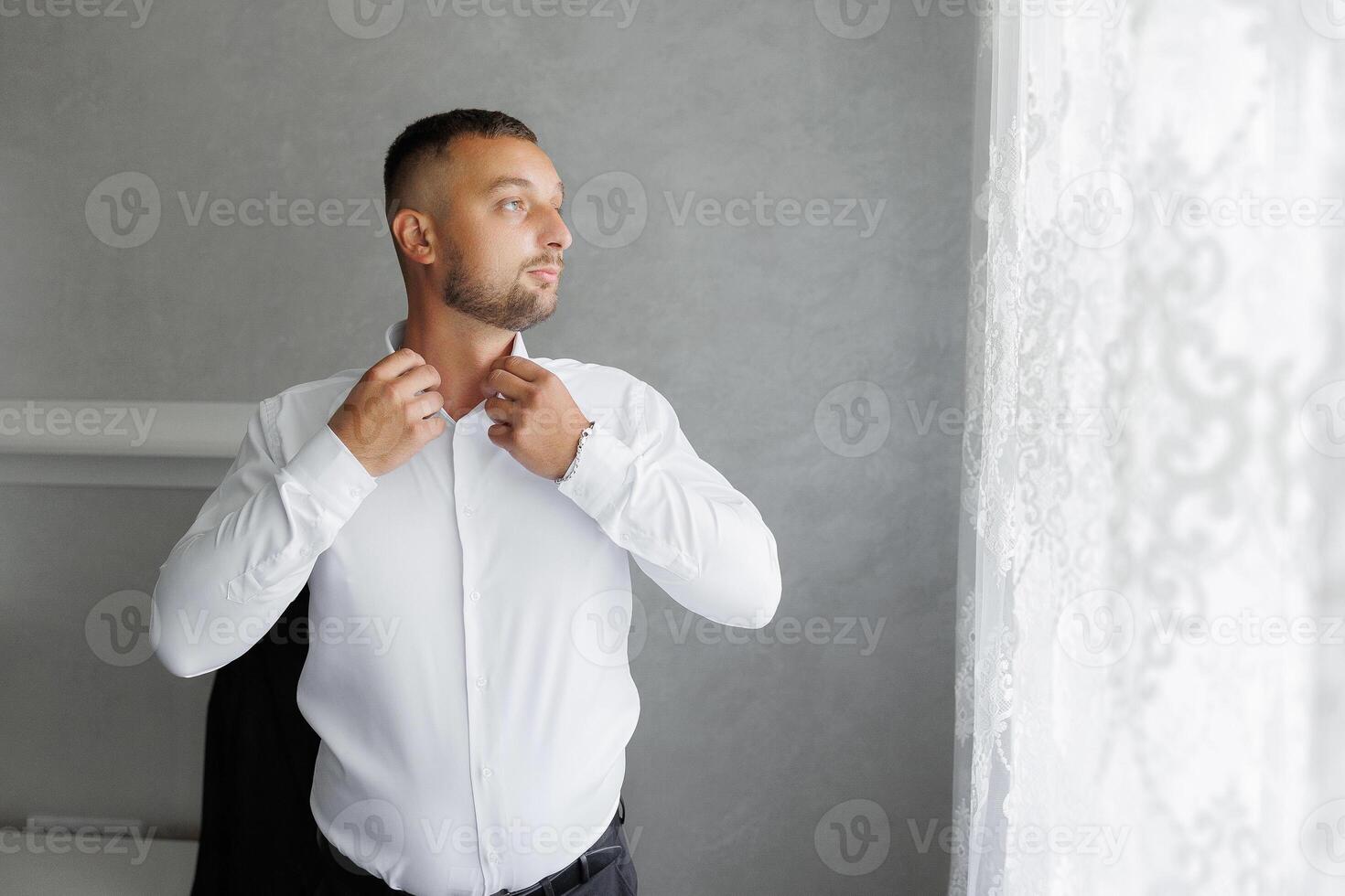 A man in a white shirt is getting dressed and looking out the window photo