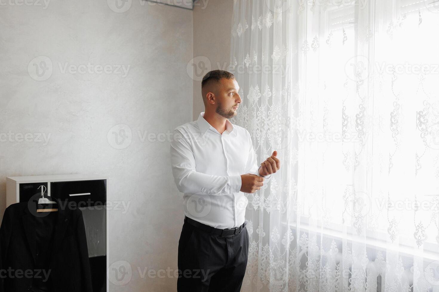 A man in a white shirt is standing in front of a window, adjusting his tie photo