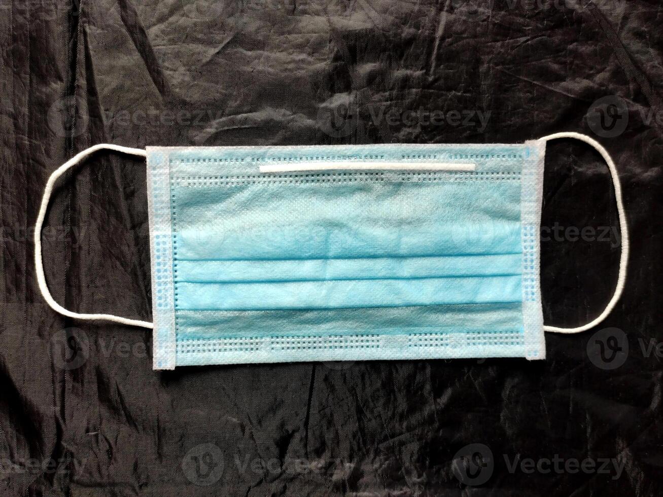 A blue disposable surgical mask with elastic ear loops and a pleated design is set against a dark background, symbolizing health, hygiene, and protection against airborne particles and droplets. photo