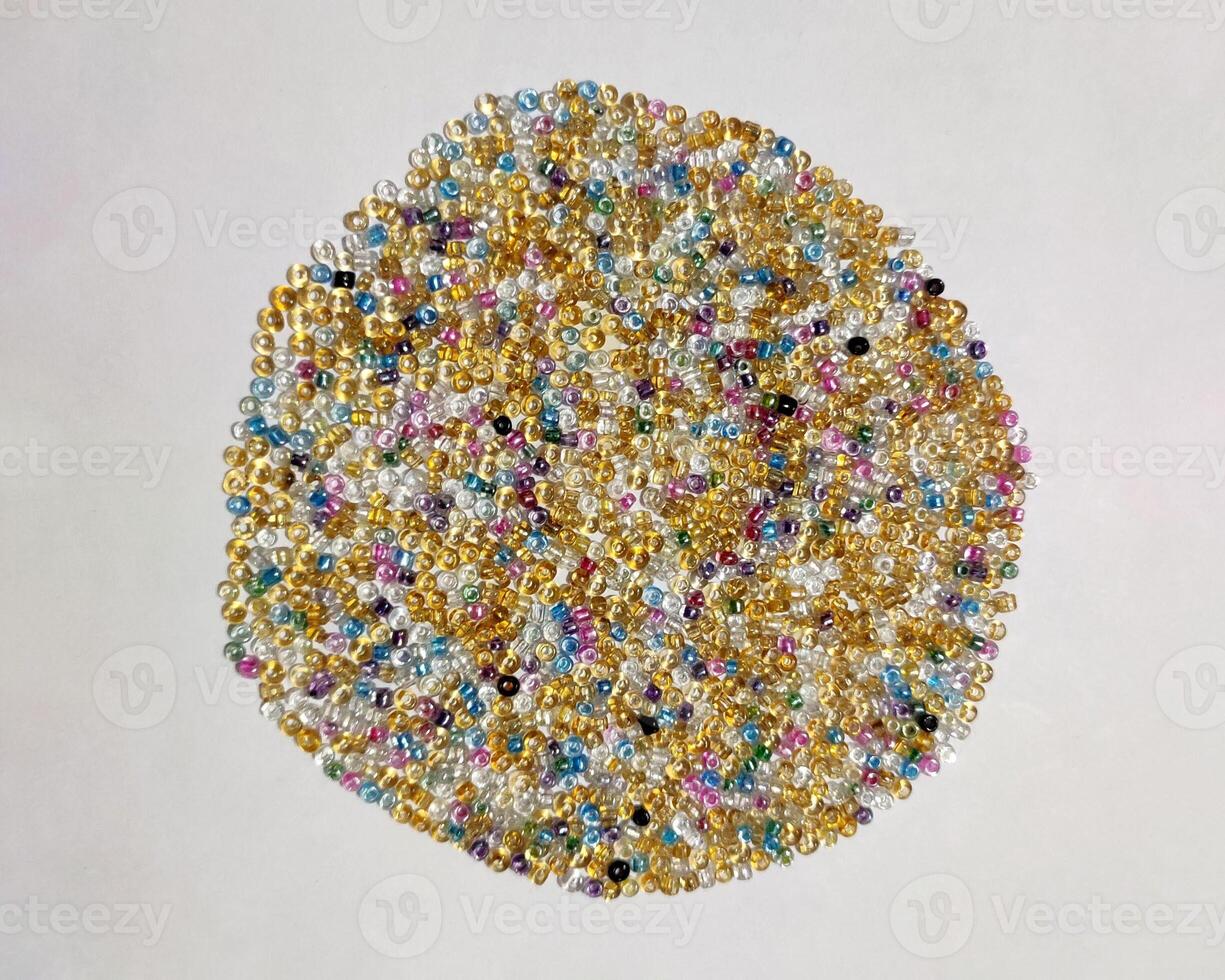 A vibrant mix of colorful seed beads in gold, silver, blue, and pink forms a circular pattern on a white background. The tiny, rounded beads with iridescent surfaces highlight crafting and creativity. photo