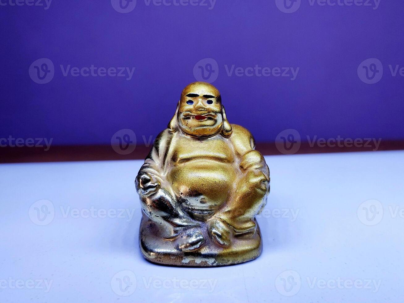 A golden Laughing Buddha figurine sits on a white surface with a purple backdrop, showcasing its joyful pose, smiling face, and plump figure, symbolizing happiness, luck, and prosperity. photo