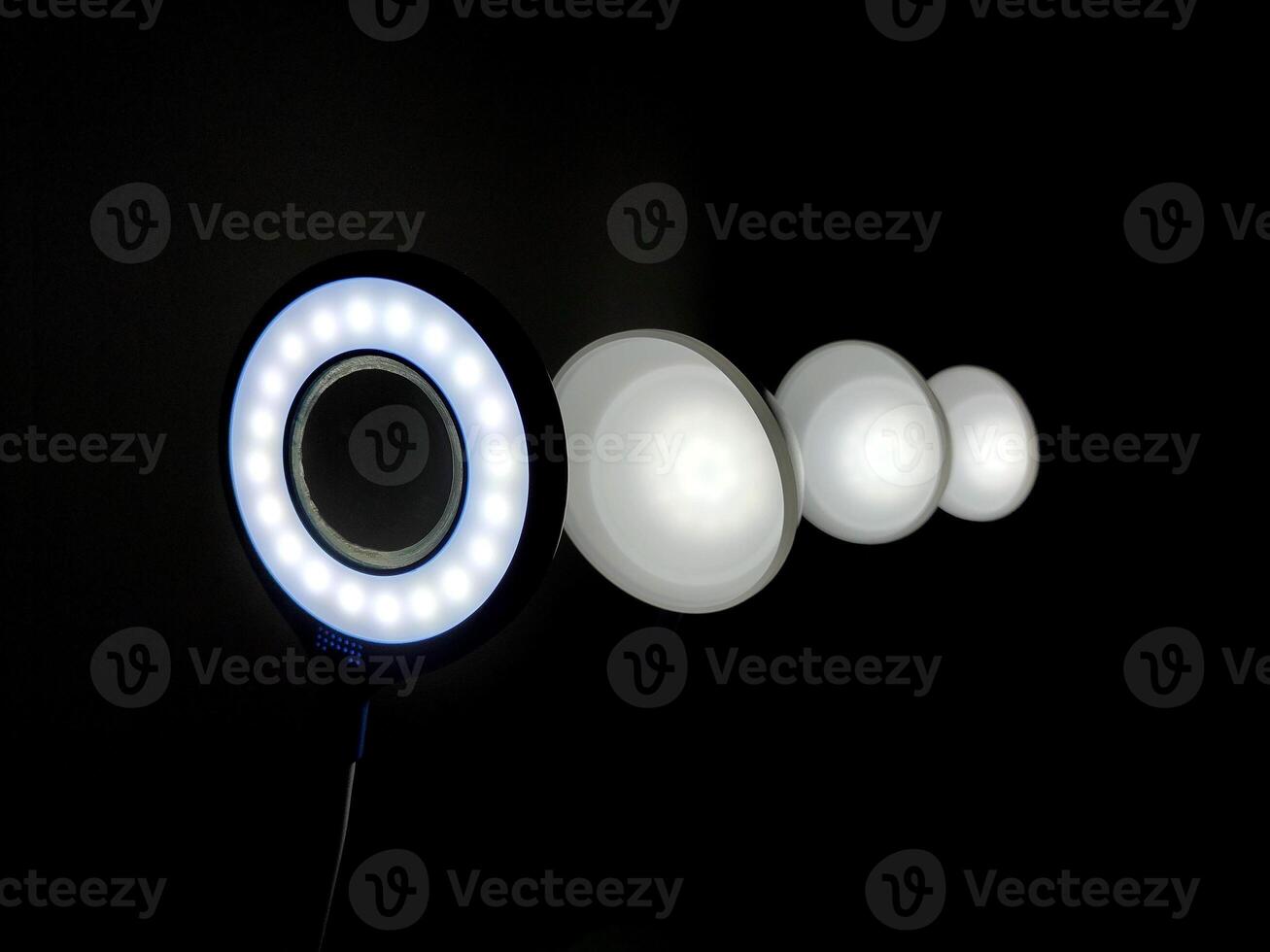 An illuminated circular LED ring light with evenly spaced bulbs shines against a dark background, accompanied by three smaller circular lights, offering a versatile setup for diffused lighting. photo