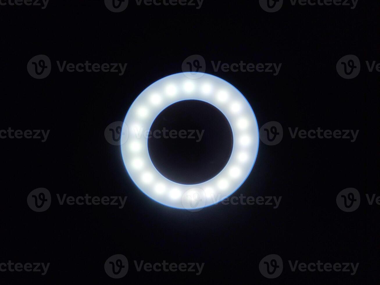 A circular LED ring light glows softly against a dark background, with evenly spaced bulbs ideal for photography, videography, or content creation. Perfect for themes of lighting, tech, or creativity. photo