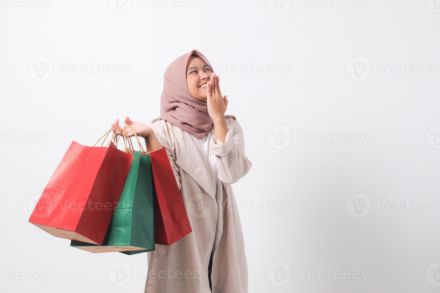 Portrait of shocked Asian hijab woman in casual suit holding paper shopping bags while covering mouth. Shopaholic girl and discount buying concept. Isolated image on white background photo