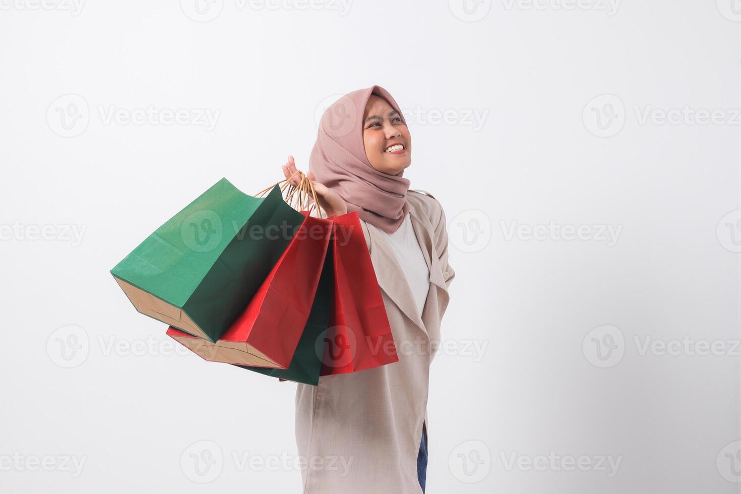 Portrait of excited Asian hijab woman in casual suit holding and showing colorful paper shopping bags. Shopaholic girl and discount buying concept. Isolated image on white background photo