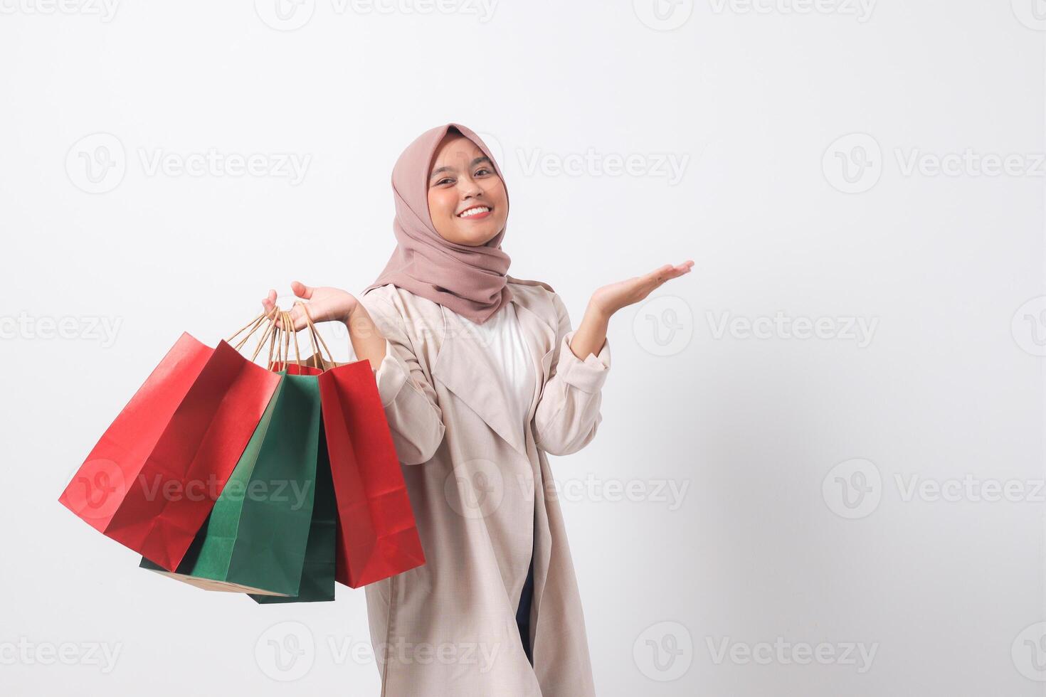 Portrait of excited Asian hijab woman in casual suit holding paper shopping bags while pointing with finger. Shopaholic girl and discount buying concept. Isolated image on white background photo