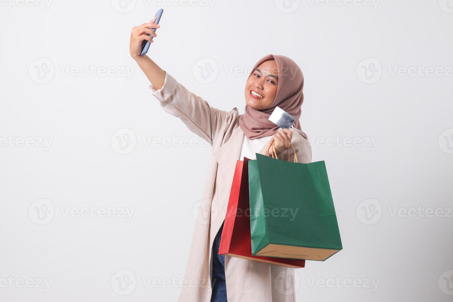 Portrait of excited Asian hijab woman in casual suit holding credit card and shopping bags while taking selfie with mobile phone. Shopaholic girl concept. Isolated image on white background photo