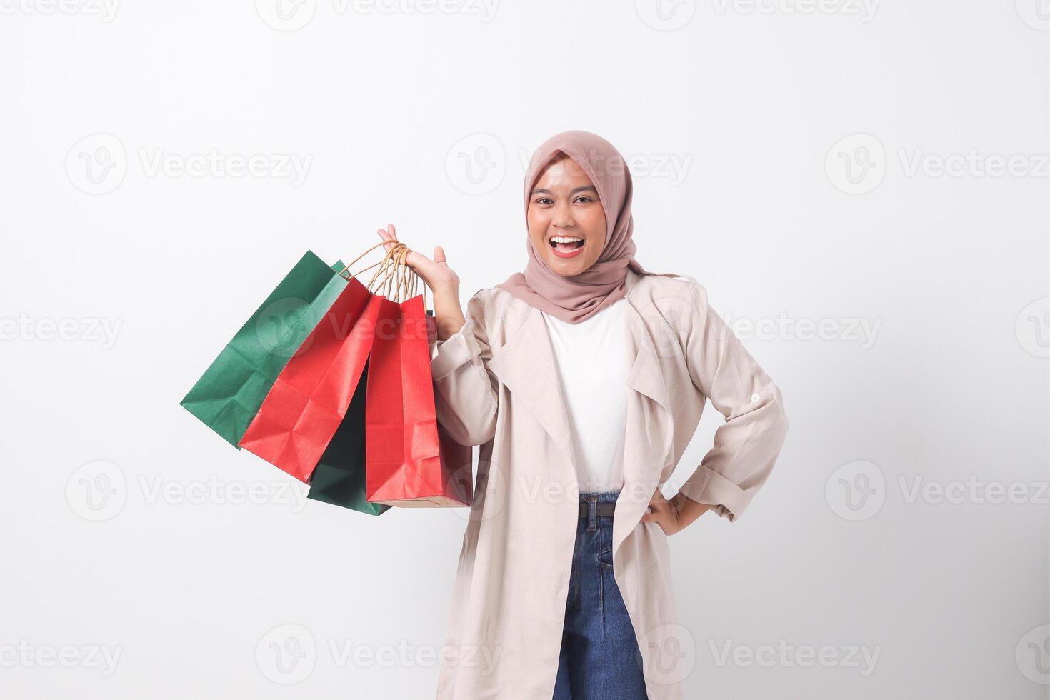 Portrait of excited Asian hijab woman in casual suit holding and showing colorful paper shopping bags. Shopaholic girl and discount buying concept. Isolated image on white background photo